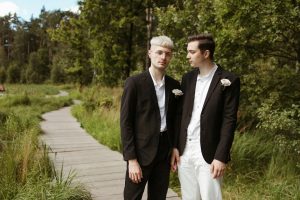 A romantic couple in black suits stands on a wooden pathway surrounded by lush greenery.