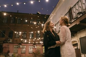 A romantic moment between two women celebrating under string lights in an urban setting at night.