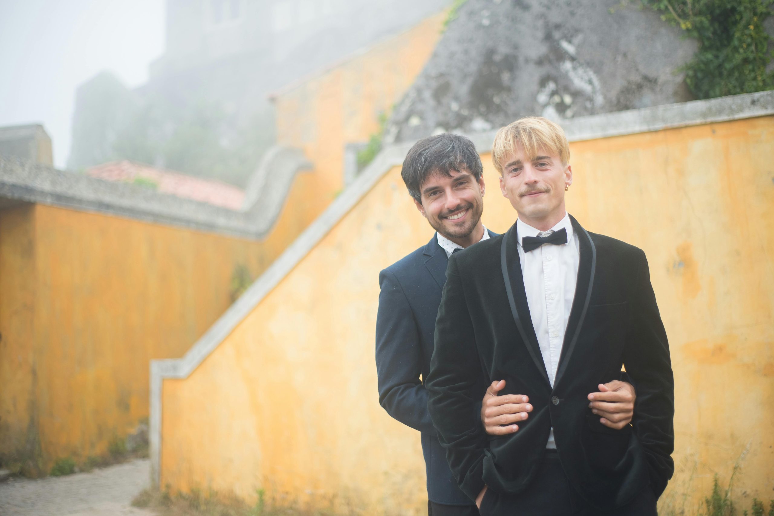 A stylish gay couple in formal attire posing outdoors against scenic walls.
